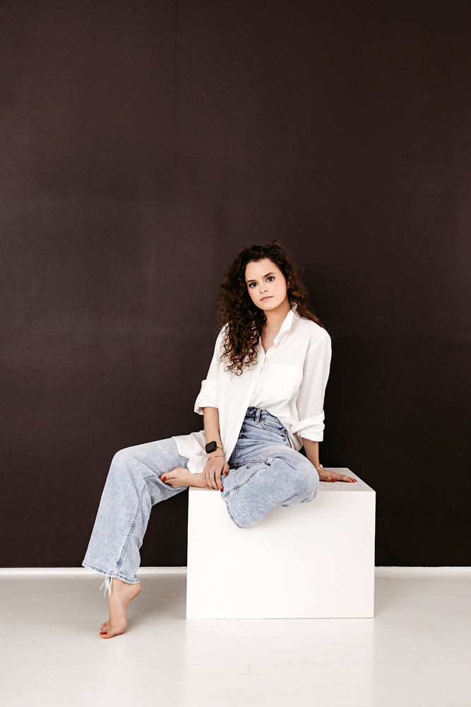 Adult woman with curly hair sitting on white cube in white button-down shirt and light jeans, dark backdrop, professional photography