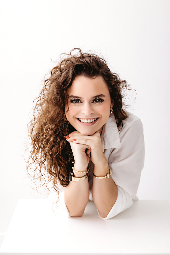 Smiling adult woman with curly brown hair in white shirt, hands under chin pose, clean white studio backdrop by professional photographer