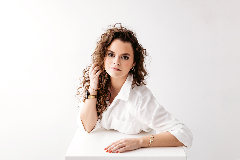 Professional young adult pictures: woman with curly brown hair in white button-up shirt posing at white table, soft studio lighting