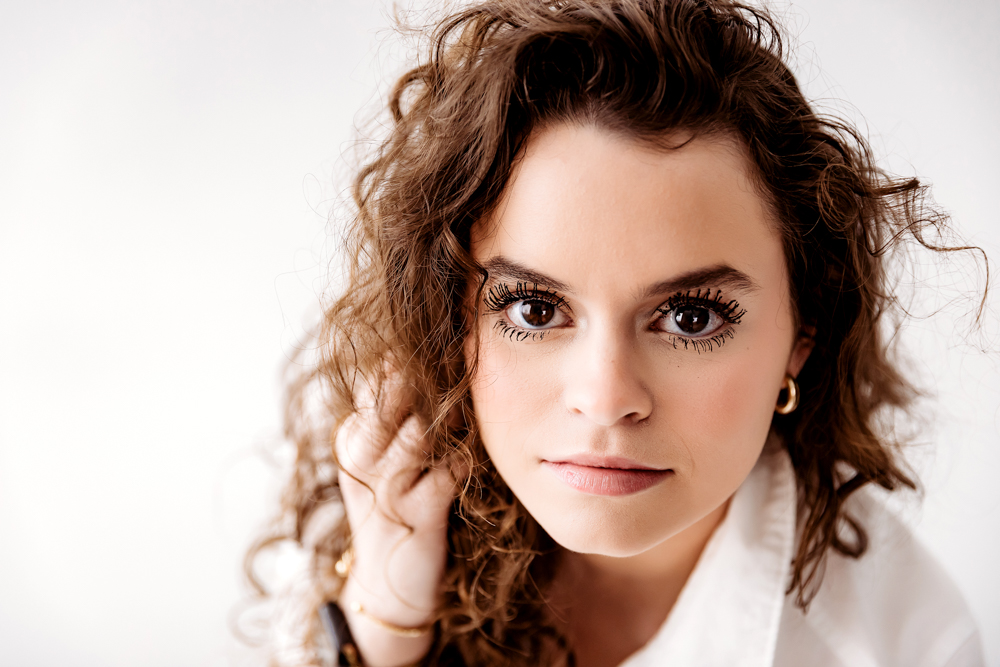 Young adult photography portrait of woman with curly brown hair in white top against soft backdrop, natural lighting
