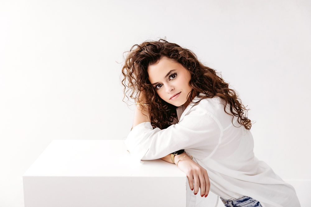 Young adult photographer with curly brown hair in white shirt posing against light backdrop, soft studio lighting