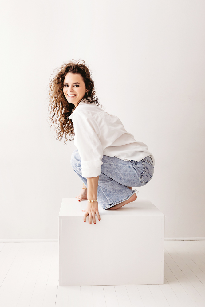 Young adult photos: smiling woman with curly brown hair in white sweater and jeans crouching on white cube, soft studio lighting
