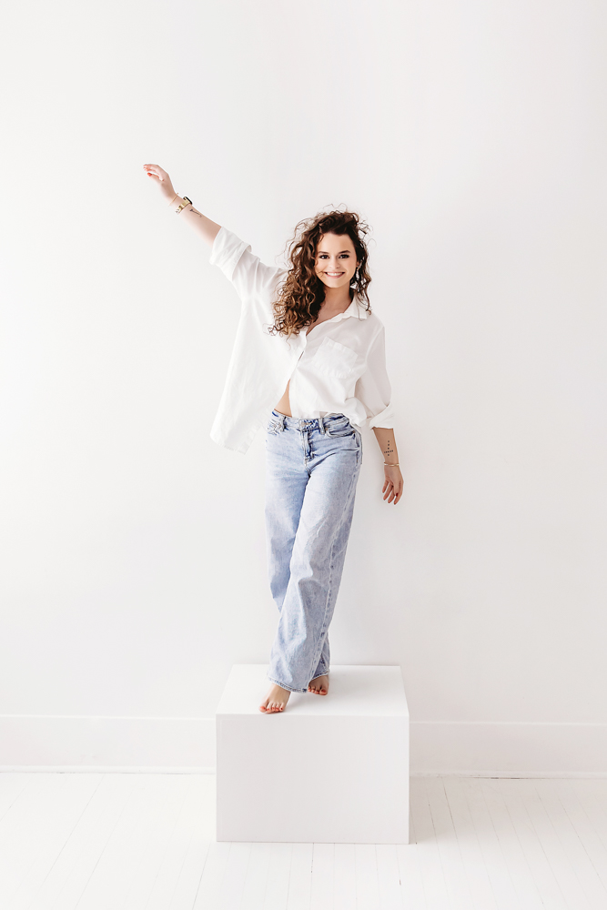 Smiling adult woman dancing in white button-up shirt and light blue jeans on white backdrop, professional photography near me