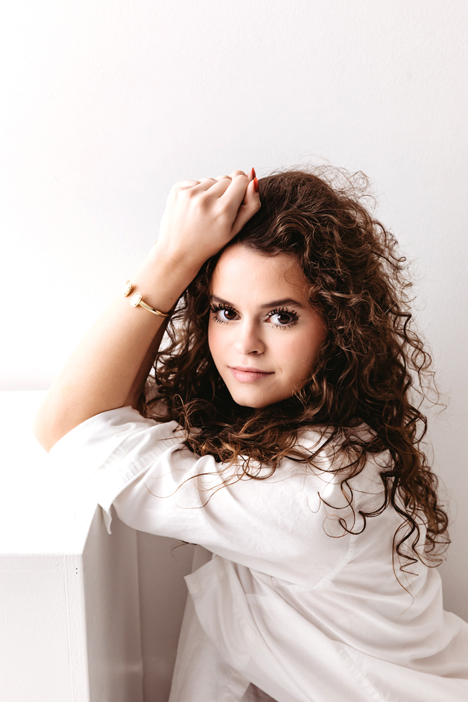 Woman with curly brown hair in white shirt posing with hand in hair against light backdrop - professional photographer near me