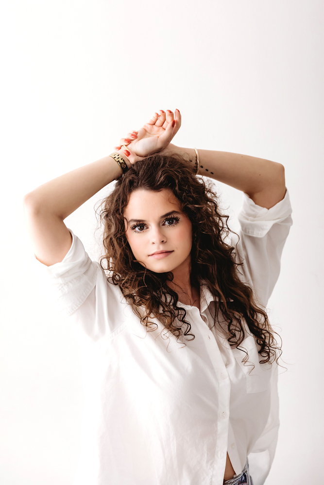 Young woman with curly brown hair in white button-up shirt posing with arms raised, professional photography studio portrait