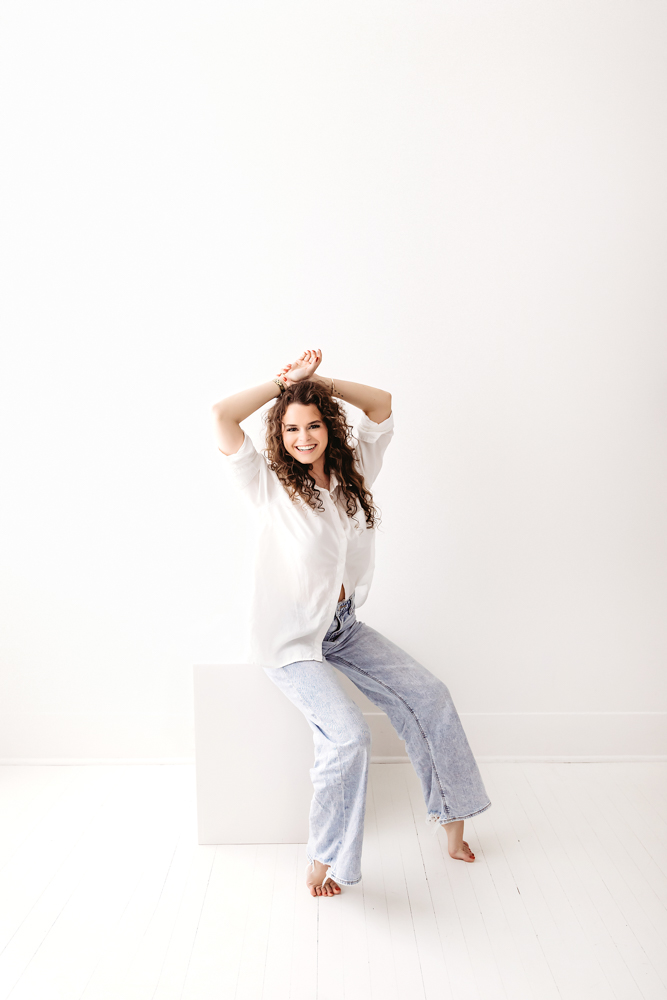 Smiling woman with curly hair in white top and jeans sitting on stool, hands in hair, professional photographer studio