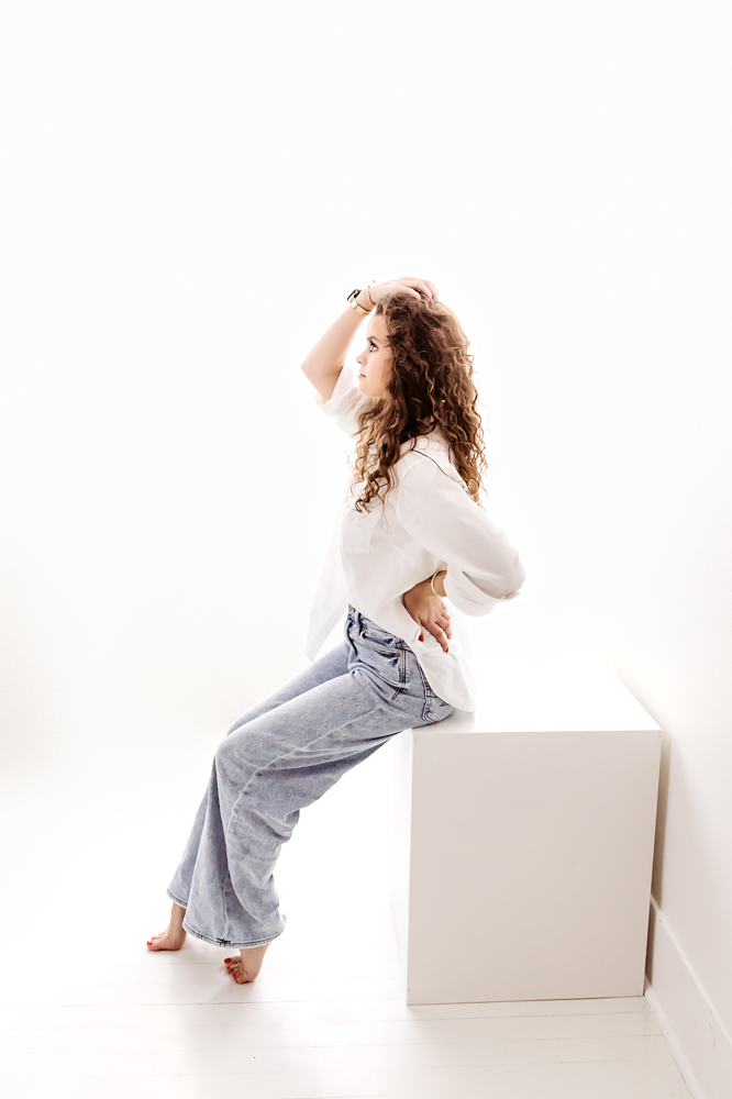 Young adult with curly hair in white shirt and jeans sitting on white cube, hand in hair, professional young adult pictures