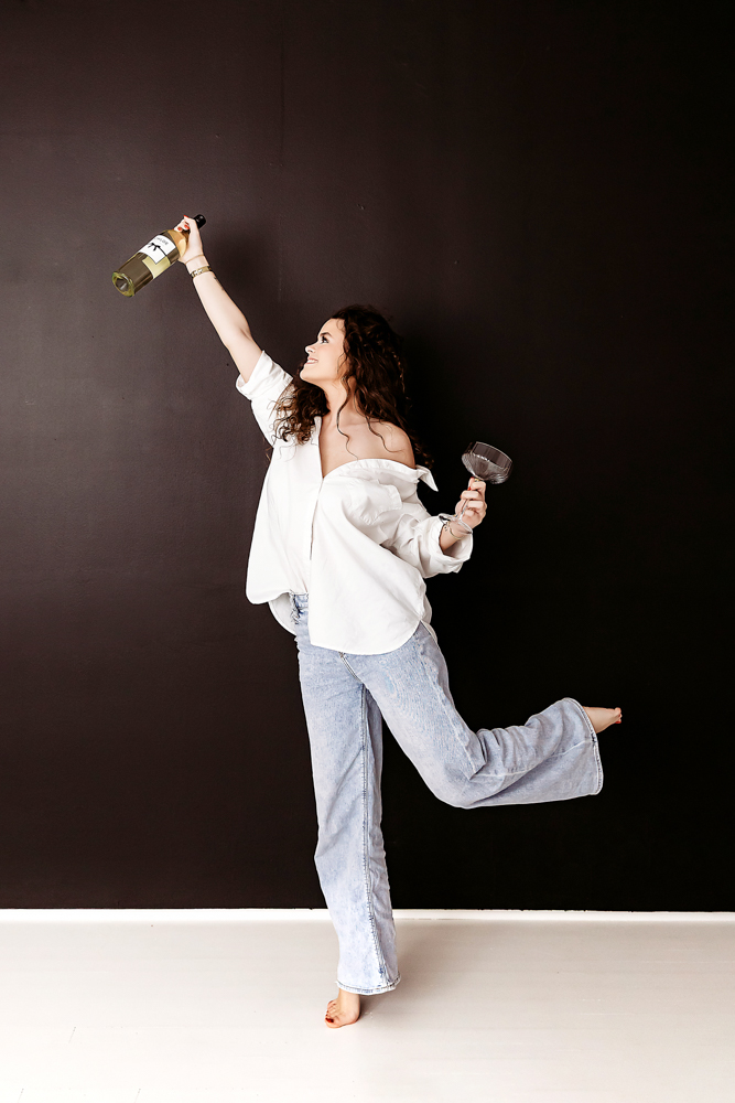Woman in white shirt and jeans dancing with wine bottle and glass against dark wall - Reflection Images by Tracy Rowell