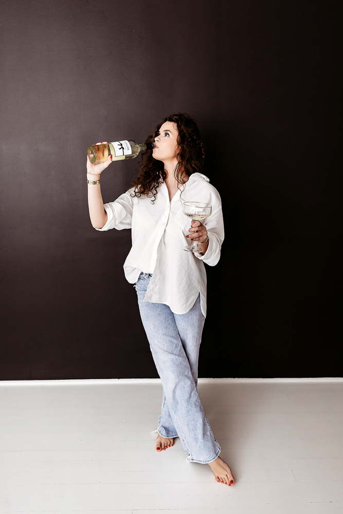 Adult woman drinking wine from bottle in white shirt and jeans against dark wall, Tracy Rowell portrait photography
