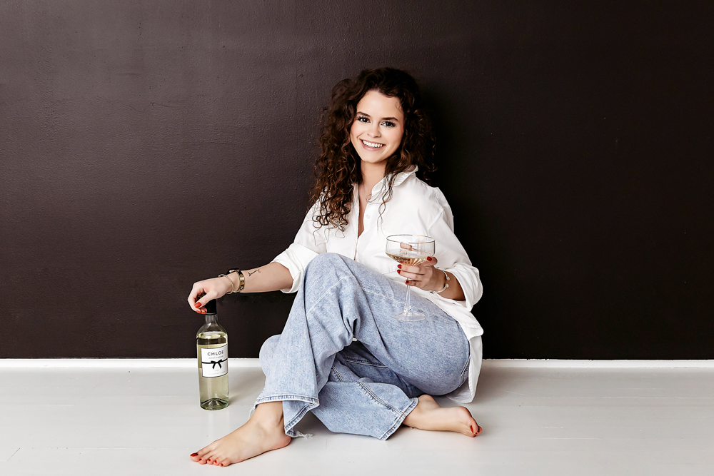 Young adult photos: Smiling woman in white shirt and light jeans sits barefoot holding wine glass against dark wall