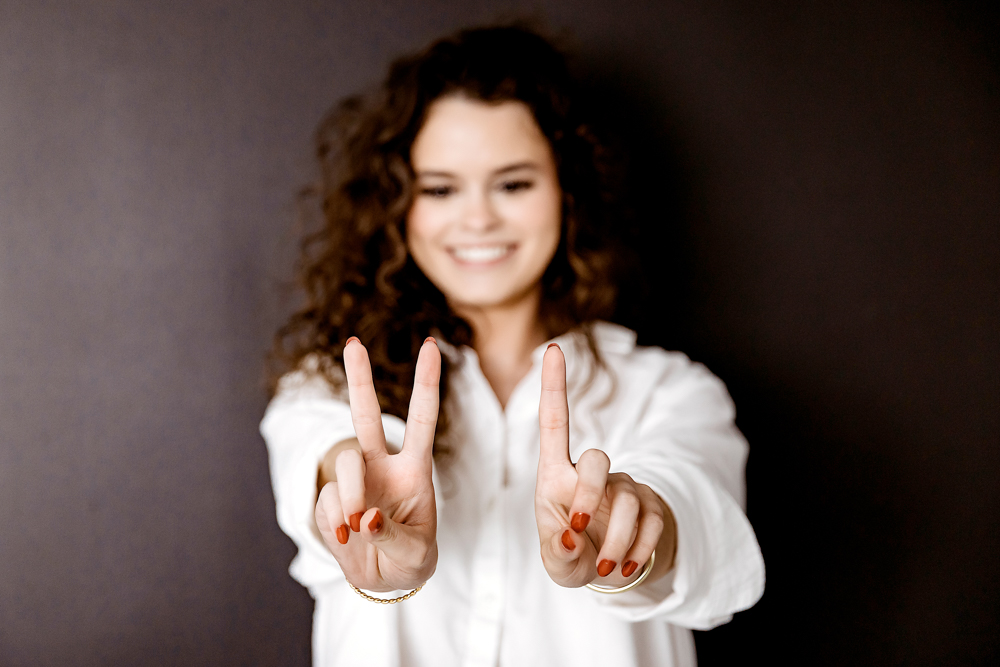 Young adult pictures: Smiling woman with curly hair making peace signs, wearing white shirt, soft lighting, gray backdrop
