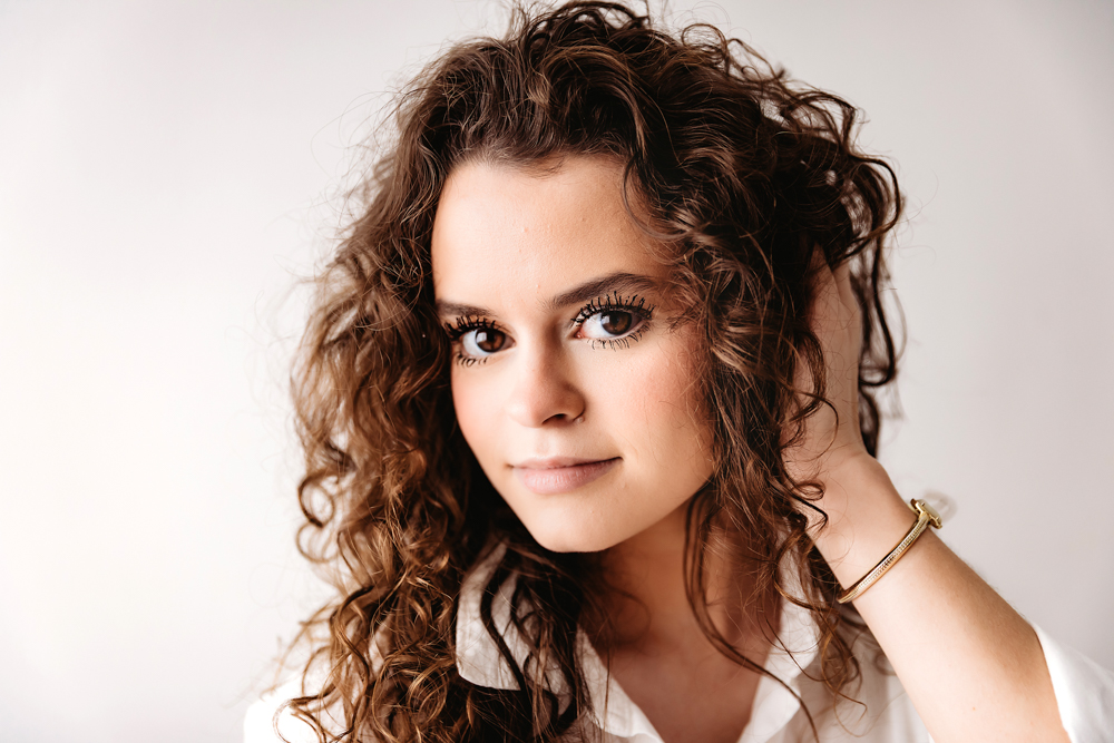 Teen with curly brown hair and gold bracelet poses against white backdrop by professional photographer