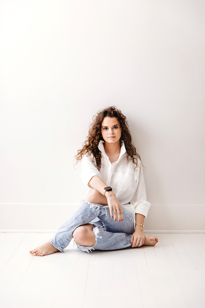 Professional young adult pictures: woman with curly hair in white shirt and distressed jeans sitting cross-legged on white floor
