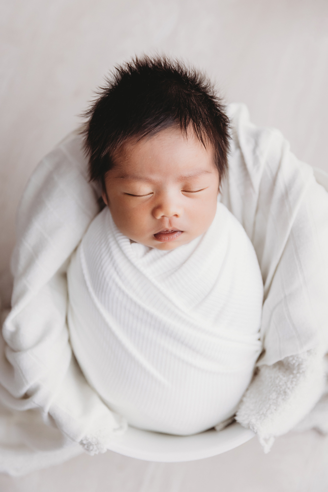 Sleeping newborn with dark hair wrapped in white blanket on cream backdrop - professional newborn pictures Florence SC