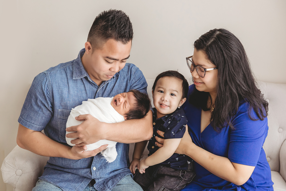Family of four on white couch - father in denim holds newborn, mother in blue top with toddler, soft studio lighting