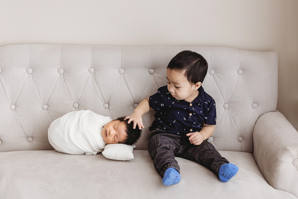 Toddler in navy patterned shirt gently touching sleeping baby in white on beige tufted sofa, professional photography near me
