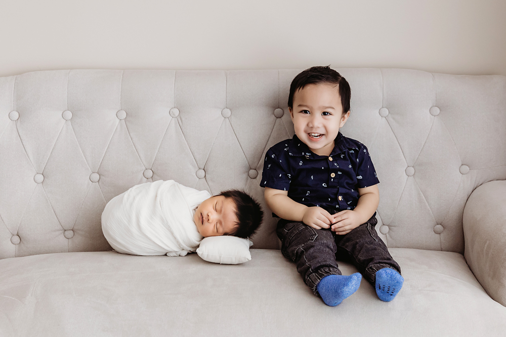 Smiling child in navy shirt sits beside swaddled newborn on tufted cream sofa, professional photographer near me portrait