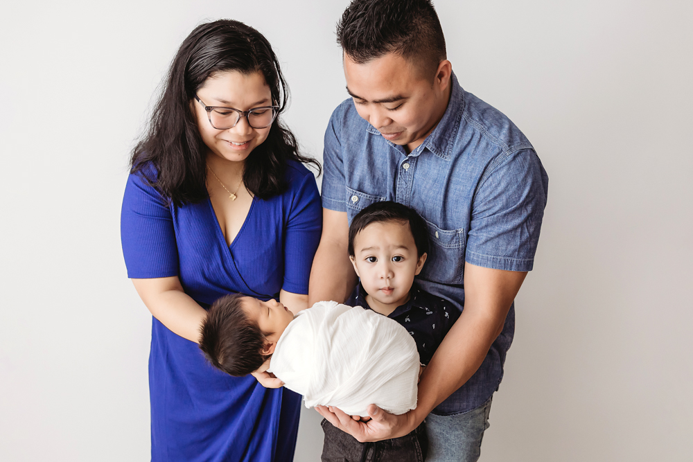 Family of four in professional photography studio: parents holding newborn in white blanket, toddler standing close on gray backdrop
