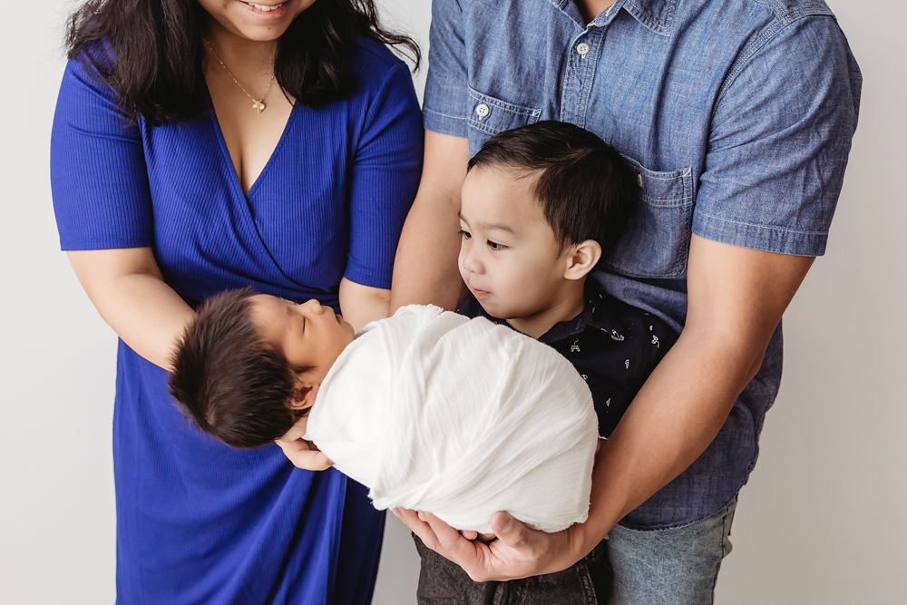 Family with newborn baby in white wrap, mother in blue dress, father in denim shirt, toddler watching, professional photography