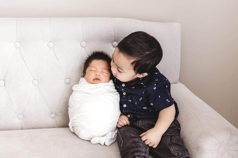 Big brother in navy shirt lovingly gazes at swaddled newborn on tufted white couch by professional photographer