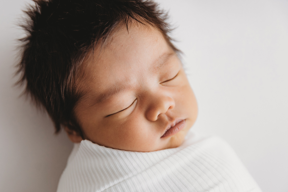 Peaceful sleeping newborn with dark hair wrapped in white blanket on cream backdrop - professional newborn pictures