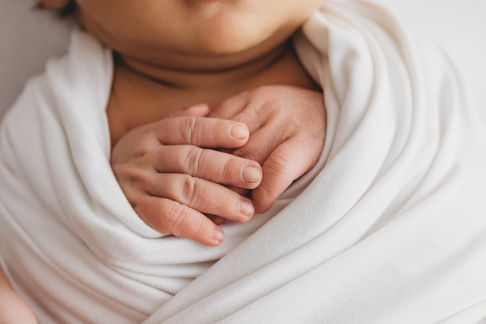 Newborn baby's tiny hands clasped together wrapped in soft white blanket, warm natural lighting, newborn photographer