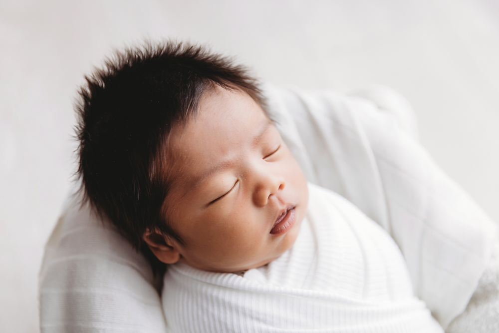 Peaceful sleeping newborn with dark hair wrapped in white knit blanket on cream backdrop, soft natural lighting, newborn photography