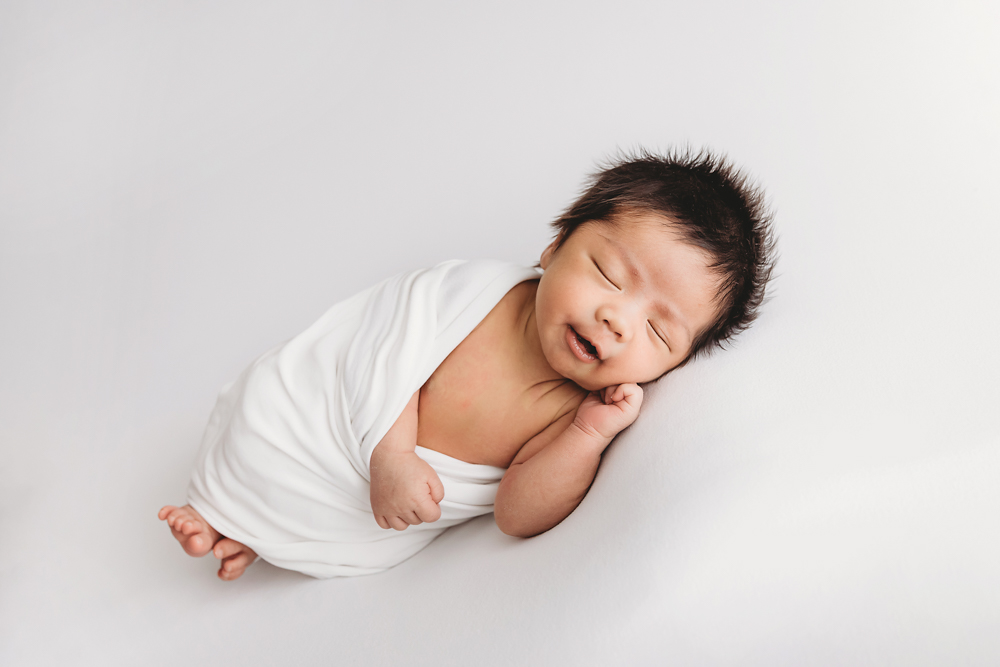Sleeping newborn baby with dark hair wrapped in white blanket on cream backdrop for newborn photos in Florence SC