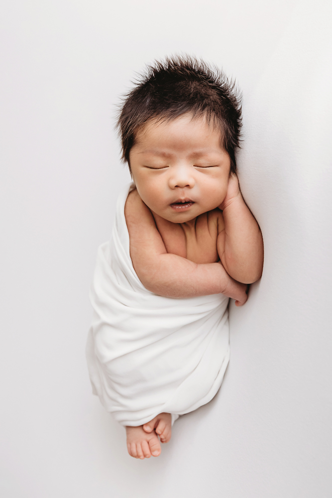 Sleeping newborn with dark hair wrapped in white blanket, hands near face on cream backdrop - newborn pictures