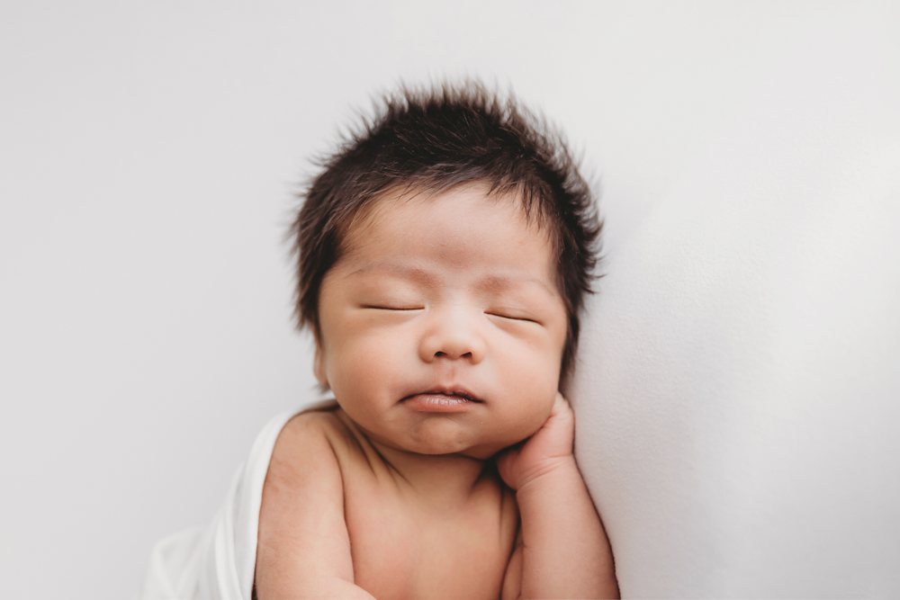 Sleeping newborn baby with dark spiky hair, hands under chin pose on white backdrop - florence sc newborn photographer
