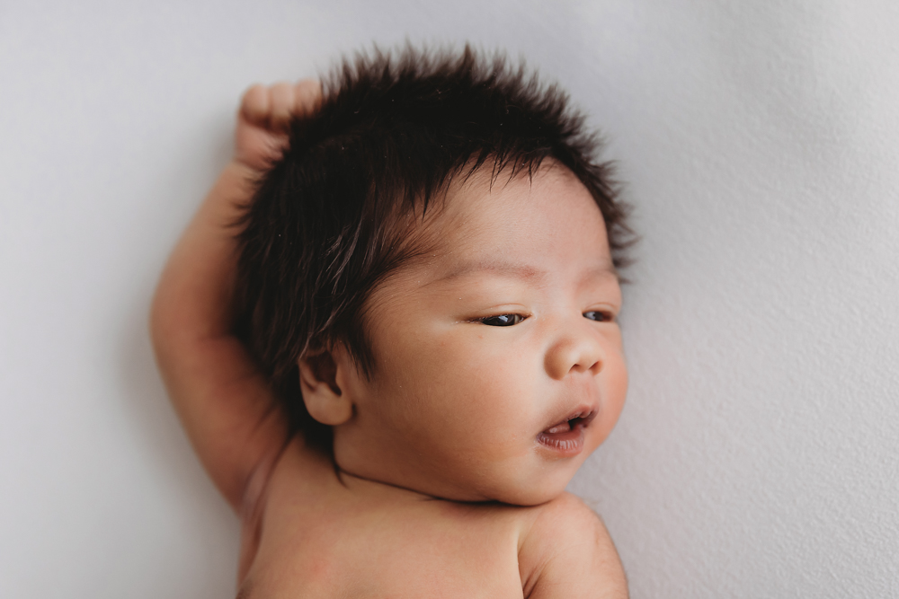 Newborn baby with dark spiky hair lying on white backdrop, hand behind head, soft natural lighting - florence sc photographer