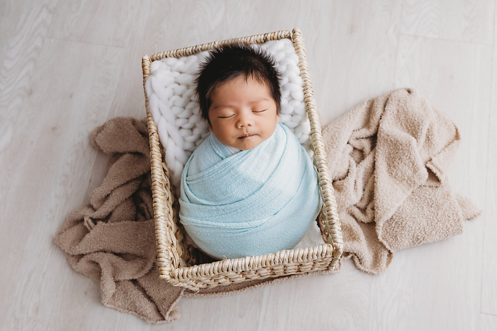 Sleeping newborn baby wrapped in soft blue blanket in wicker basket with beige textiles, professional photography studio near me