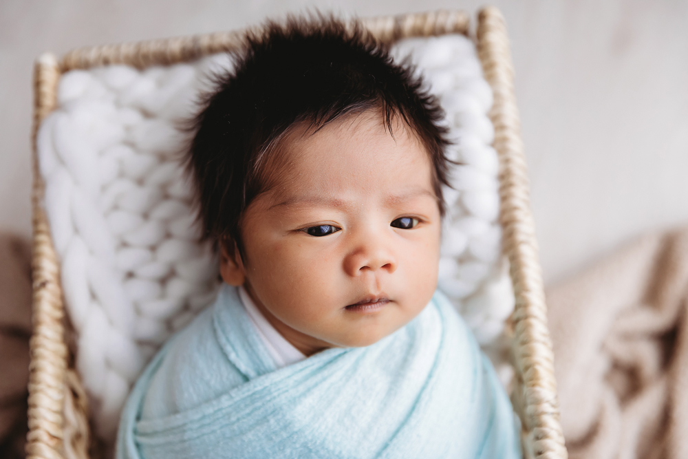 Alert newborn baby with dark hair in soft blue swaddle lying in wicker basket with white blanket, professional photography near me