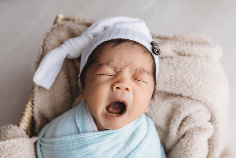 Yawning newborn in white knit hat and blue wrap on cream blankets, professional photographer near me portrait