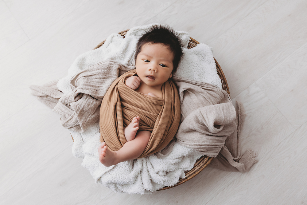 Alert newborn baby in tan wrap lying in wicker basket with cream blankets in professional photography studio