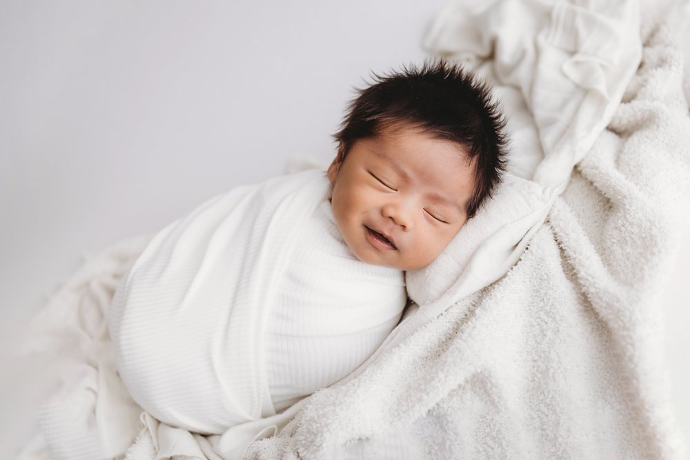 Sleeping newborn with dark hair wrapped in white knit blanket on cream backdrop by newborn photographer in Florence SC