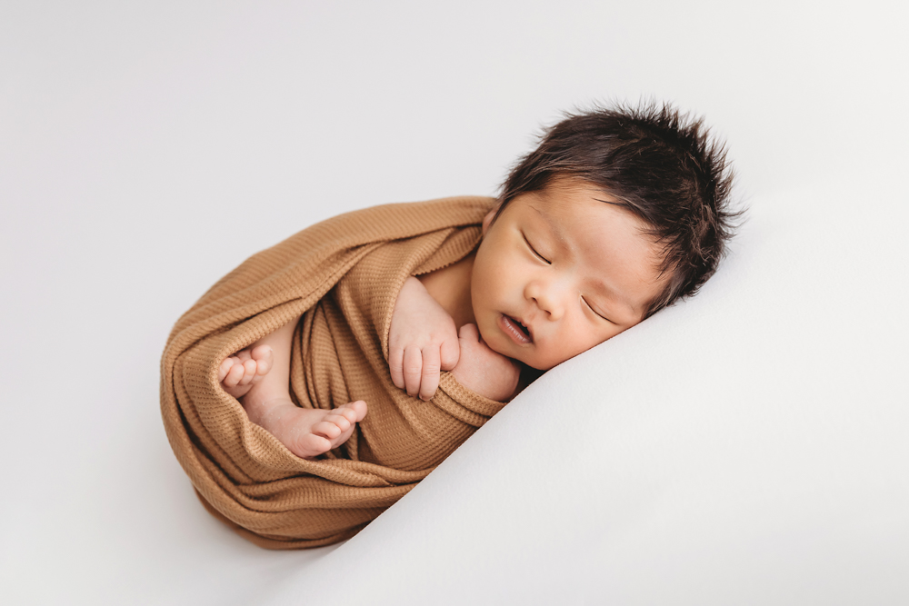 Sleeping newborn baby wrapped in camel-colored knit blanket on white backdrop by professional photographer