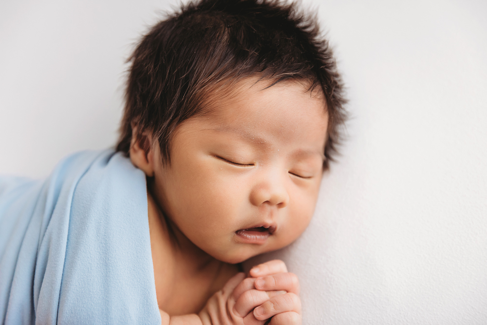 Sleeping newborn with dark hair wrapped in soft blue blanket on white backdrop - professional newborn pictures