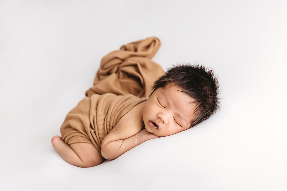 Sleeping newborn wrapped in camel-colored knit swaddle on white backdrop, peaceful newborn photography with soft lighting
