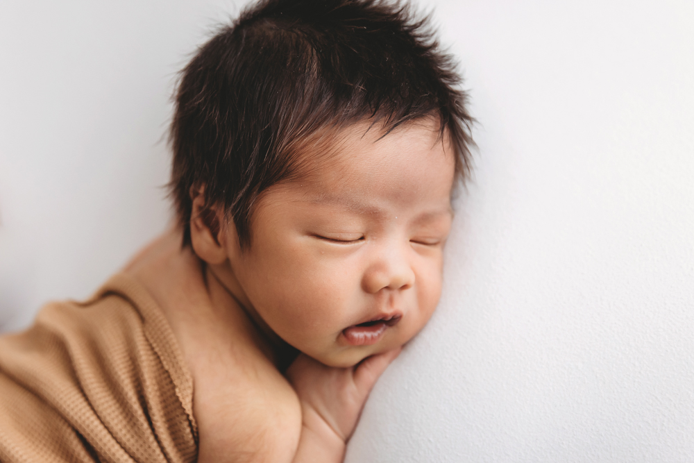 Sleeping newborn with dark hair wrapped in beige blanket on white backdrop by Florence SC newborn photographer