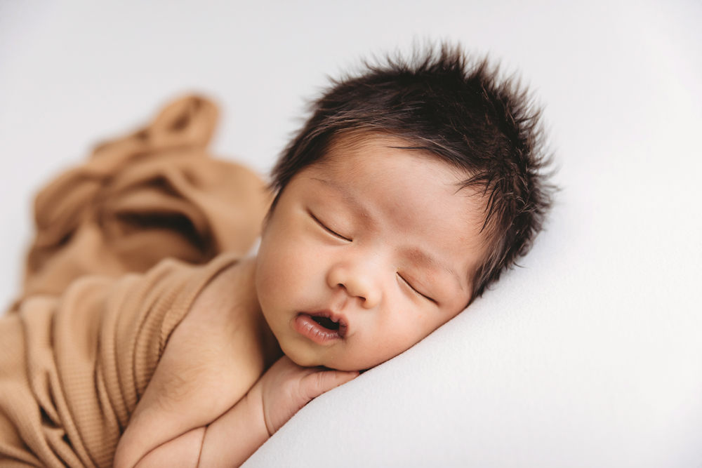 Sleeping newborn baby with dark hair wrapped in tan knit blanket on white backdrop - Reflection Images by Tracy Rowell