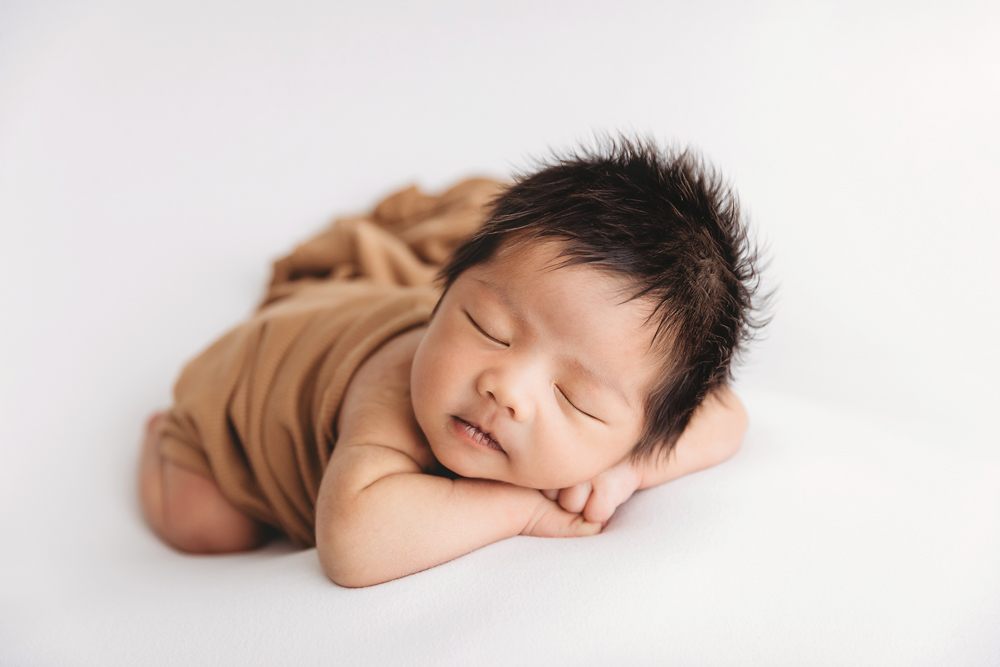Sleeping newborn baby with dark hair wrapped in tan blanket on white backdrop, soft lighting by Tracy Rowell