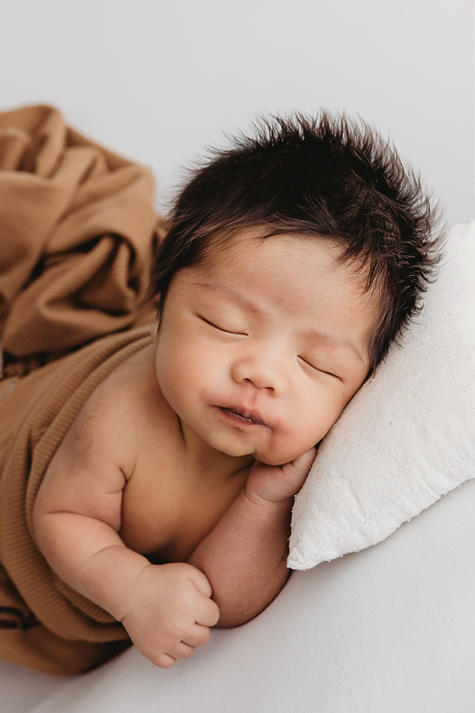 Peaceful sleeping baby with dark spiky hair wrapped in camel brown blanket on white backdrop for newborn photos