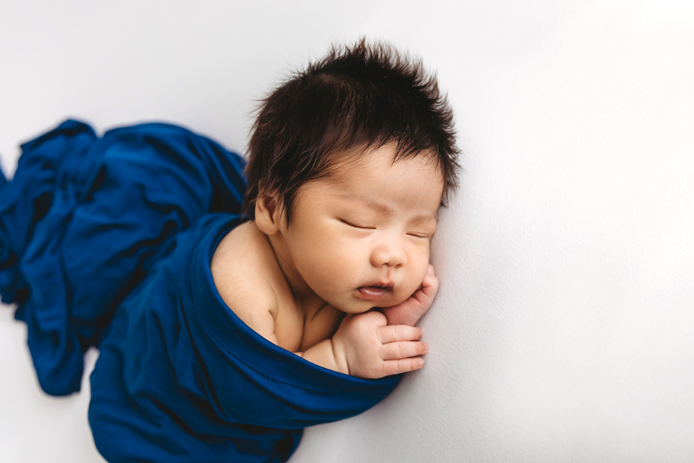 Sleeping newborn baby wrapped in royal blue fabric on white backdrop for professional newborn pictures session