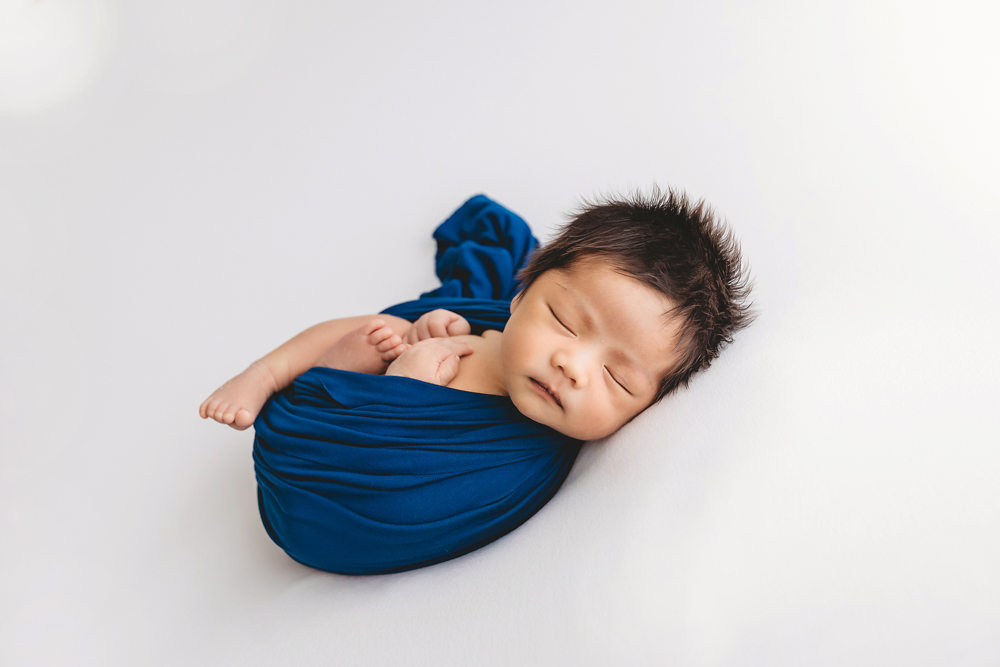 Sleeping newborn wrapped in blue fabric with matching headband on white backdrop, florence sc newborn photographer portrait