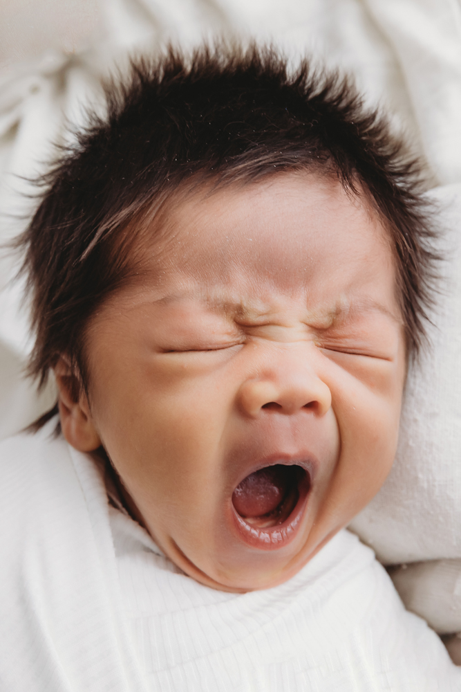 Crying newborn baby with dark hair in white fur-trimmed hat on cream blanket - Reflection Images by Tracy Rowell