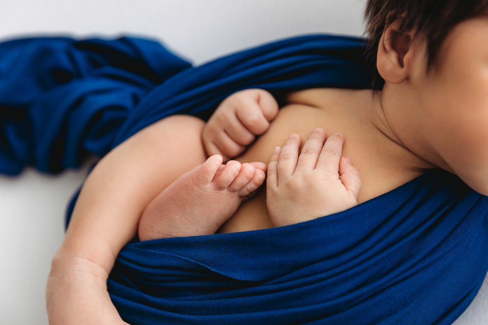 Newborn baby nestled against mother in blue wrap, tiny hands and feet visible, soft lighting professional photography studio near me