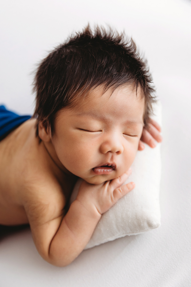 Sleeping newborn with dark spiky hair on white blanket, soft natural lighting, professional photography near me