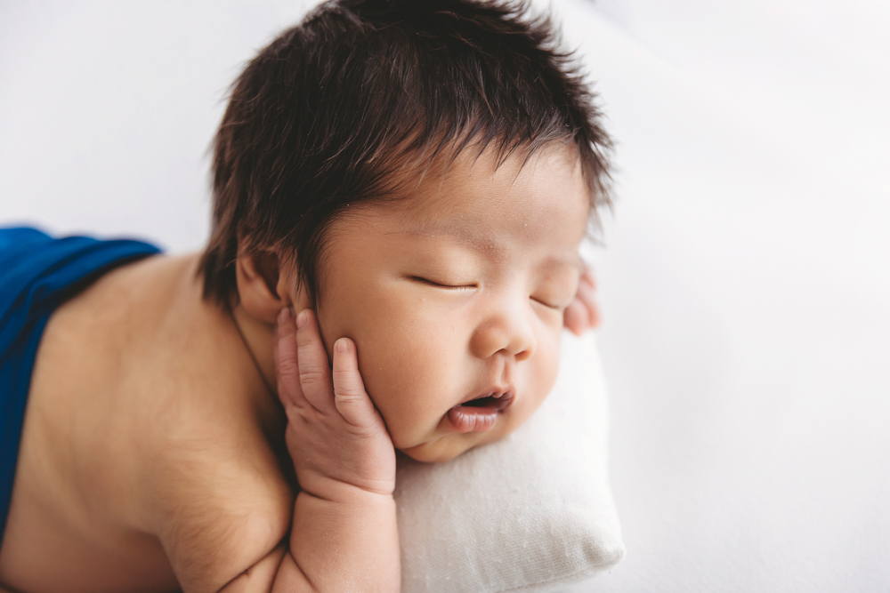 Sleeping newborn baby with dark hair in blue wrap, hand by face on white backdrop in professional photography studio