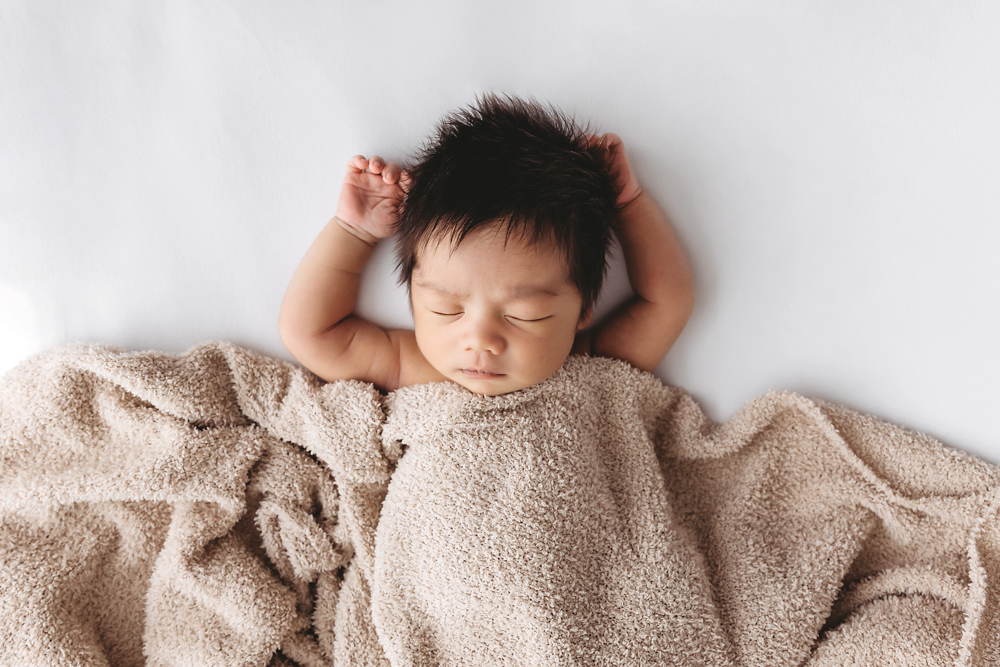 Sleeping newborn baby with dark hair and arms raised, wrapped in beige textured blanket on white backdrop, professional photography